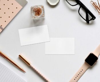 business card lying flat lay surrounded by a glass, pink smart watch, pink pens and pink notebook.
